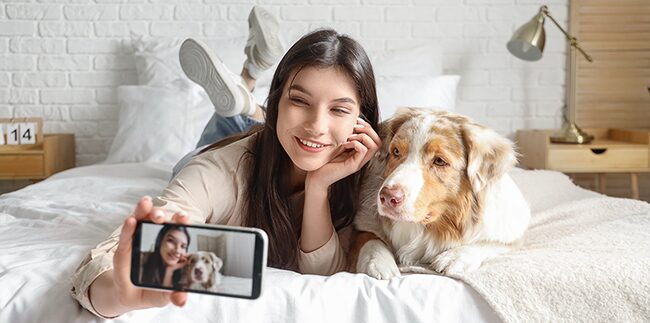 A woman and a red merle Australian Shepherd laying side-by-side on a bed taking a selfie with a smart phone.