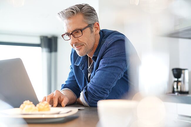 A grey-haired white man in a casual blue button down shirt sitting at his kitchen table looking at his laptop intently.