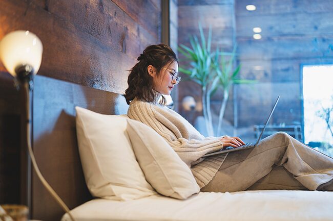 An Asian woman sitting on a bed, propped up with pillows, with her laptop open on her lap typing.