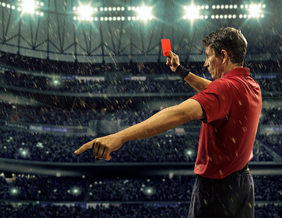 A soccer referee in a red jersey holds up a red card and is pointing to the left. He's in a stadium with bright lights and the outlines of fans in the stands