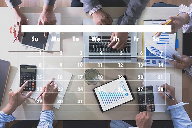 Overhead view of a table with 5 people's hands doing things: 1 has a tablet, 1 has a laptop, 1 had a clipboard with papers and holding a pencil, 1 has a notebook and pen with a calculator and the other also has a calculator, pencil and notepad. There's a white calendar outline overlaid over the image.